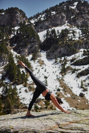 Young female doing downward dog yoga pose with one leg up in mountain scenery