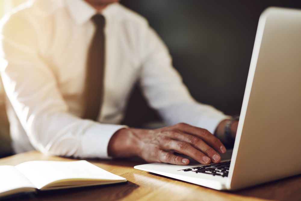 Man in shirt and tie typing on laptop with notebook