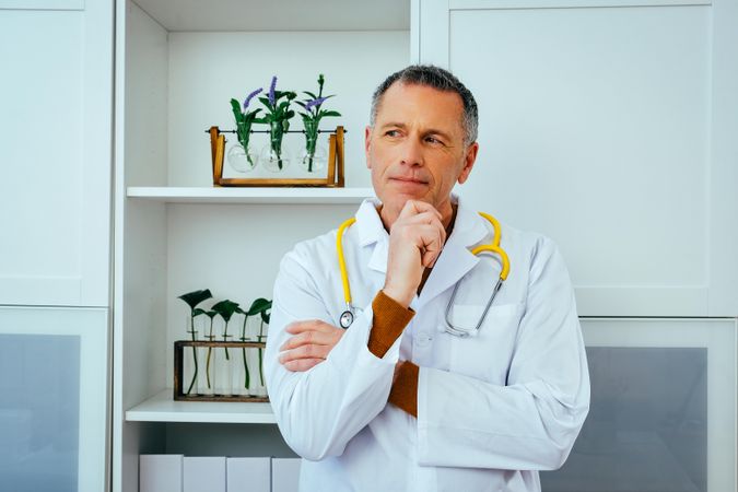 Thoughtful mature doctor standing in a clinical setting with hand to his chin, looking away