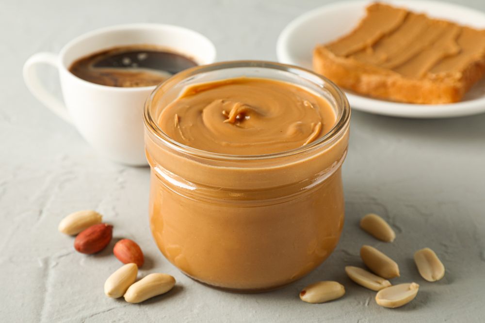Glass jar with peanut butter, peanut, cup of coffee and peanut butter sandwich on grey background, closeup and space for text