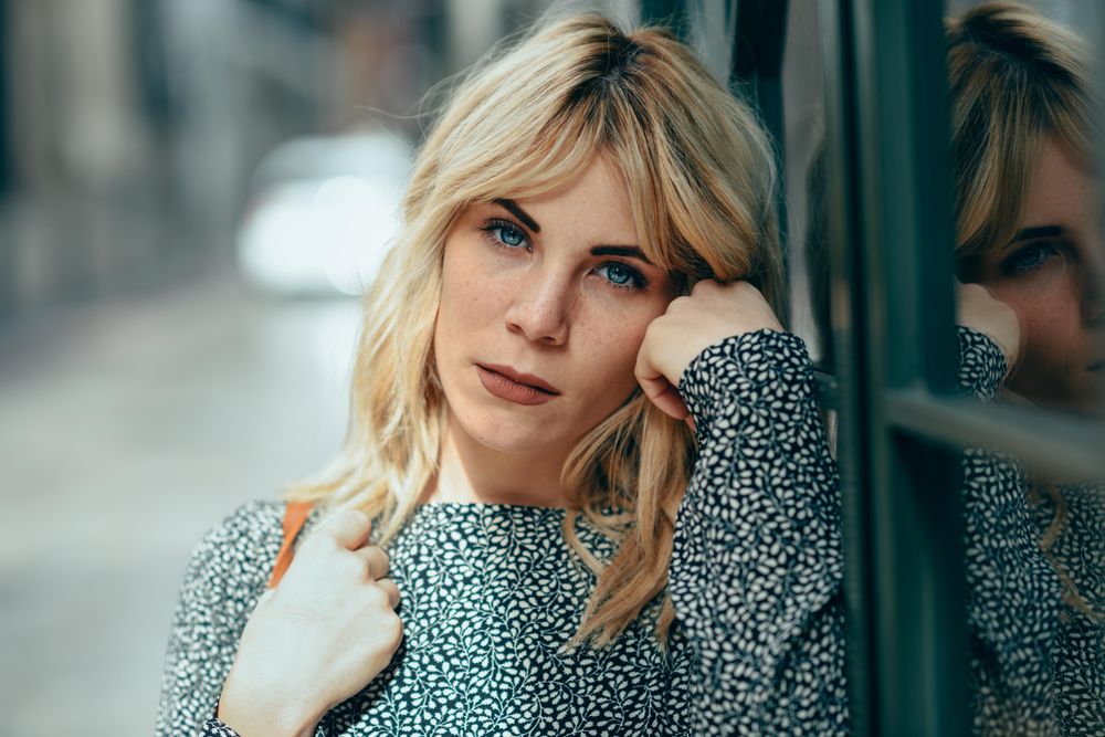 Close up of blonde female wearing casual clothes standing next to windows outside, copy space