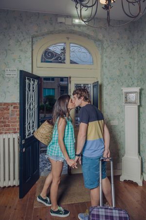Couple kiss in entrance of home with roller suitcase