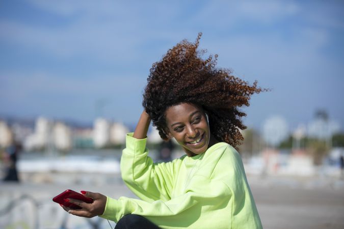 Smiling female with curly hair in bright green shirt standing listening to music on dock