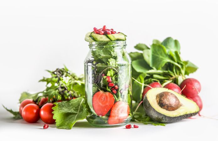 Green salad in jar with ingredients: green lettuce leaves, tomato, strawberry, avocado, cucumber and pomegranate seeds