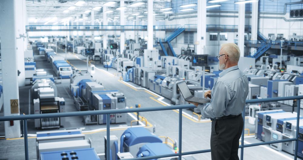 Engineer Standing on a Platform, Using Laptop Computer at an Electronics Factory. Machines are Undergoing Maintenance, Specialist Monitoring the Progress Through Online Software