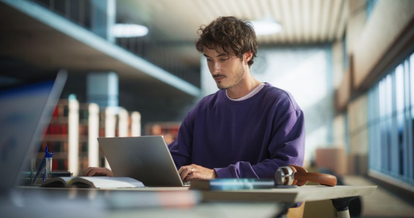Portrait of a Stylish Male Student Working on His University Exercise on a Laptop Computer. Young Handsome Man Studying an Online College Course in an Empty Quiet Public Library