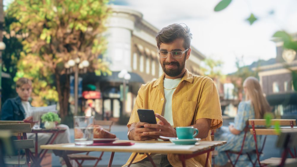 Handsome Indian Man Sitting on a Terrace in a Cafe, Having a Cup of Coffee with Pastry.