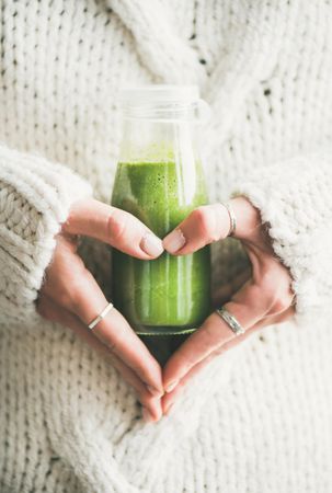Woman’s hand making heart shape, holding green smoothie wearing sweater