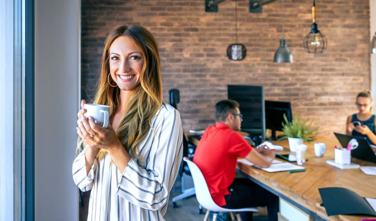 Happy businesswoman looking at camera drinking coffee in the office