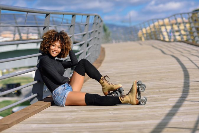 Smiling woman with afro hairstyle resting on wooden bridge in roller skates