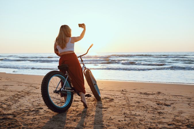Cheerful woman taking selfie on smartphone while resting with bicycle at sunny shore