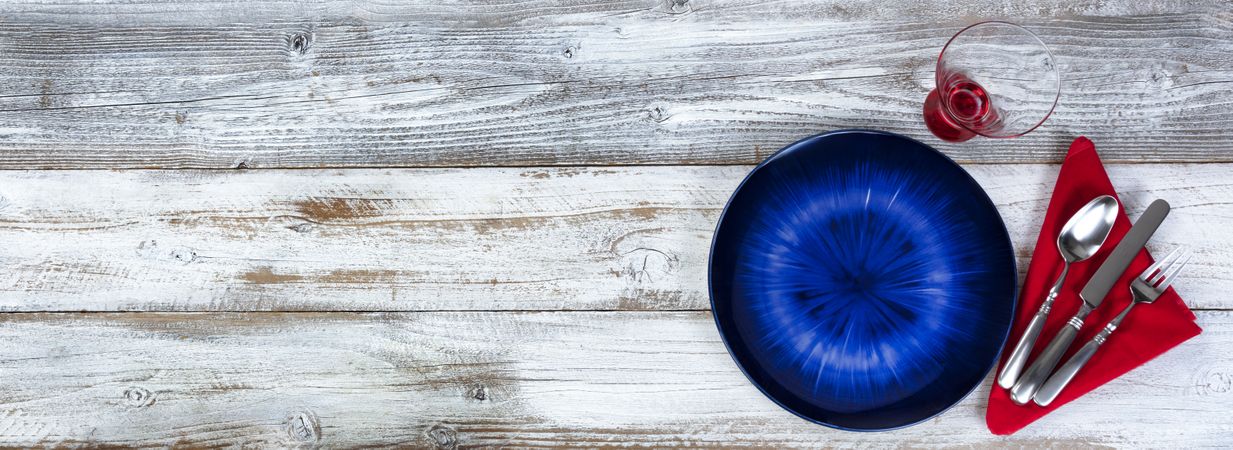 Table setting for Valentines Day celebration in flat lay format with blue plate, red napkin, silverware and drinking glass on rustic wood
