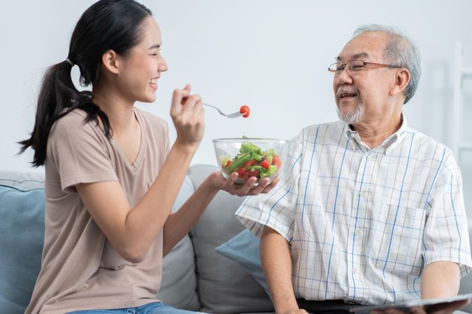 Female feeding older male salad at home while he uses tablet