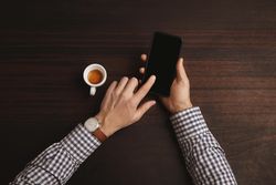 Man wearing plaid shirt and leather watch scrolling on phone with espresso shot on table