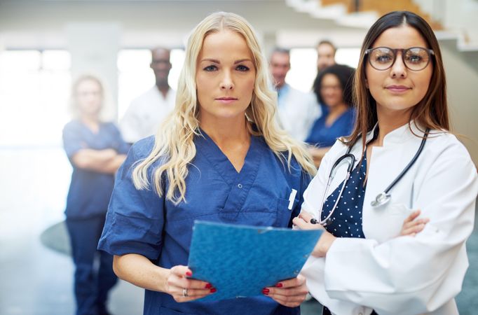 Two women working in the medical field pictured in scrubs, lap coat and stethoscope