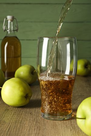 Glass and bottle with apple cider and green apples on wooden table on green wooden background