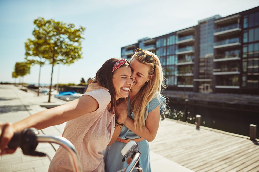 Two friends with arms on each others shoulders while walking with bike next to river, copy space
