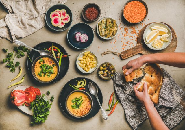 Spread of yellow lentil soup bowls, with bread, vegetable garnishes with man breaking bread