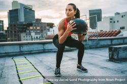 Woman doing squats holding a medicine ball with an agility ladder by her side on rooftop bEAnAb