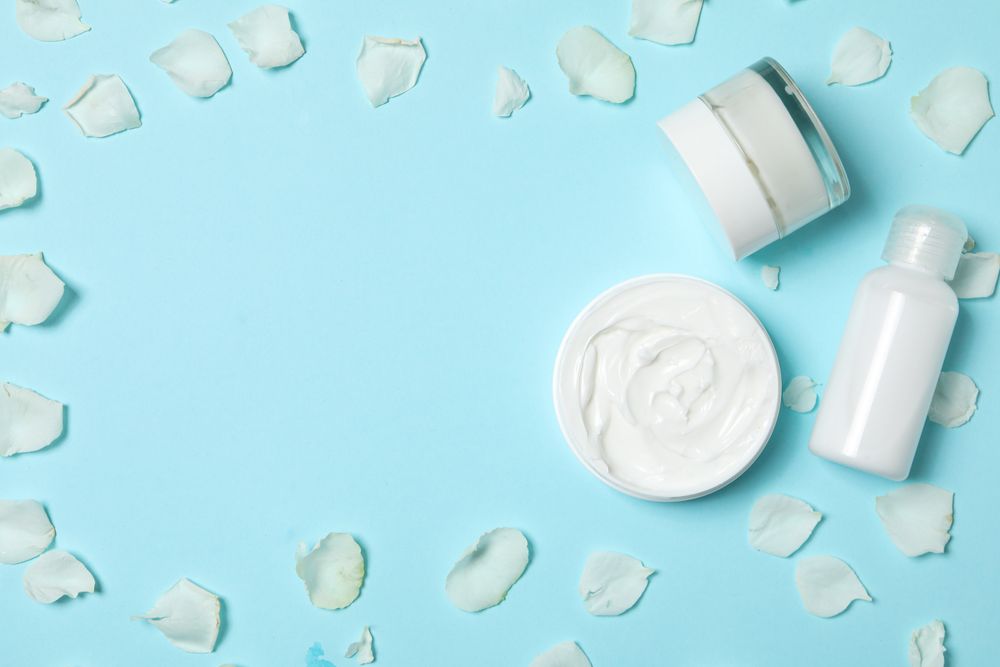 Jars with cream and rose petals on blue background, space for text