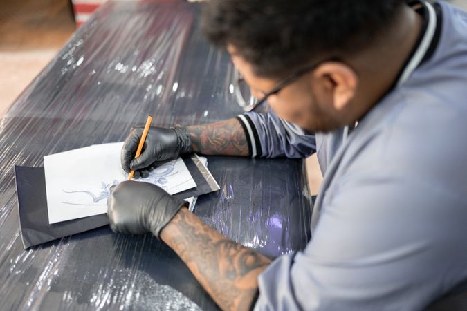 A tattoo artist is drawing a design stencil on his work stretcher before tattooing