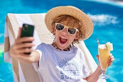 Cheerful boy holding drink and using smartphone on sunbed
