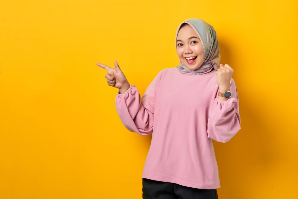 Happy Muslim woman in headscarf pointing to space beside her and raising fist in celebration