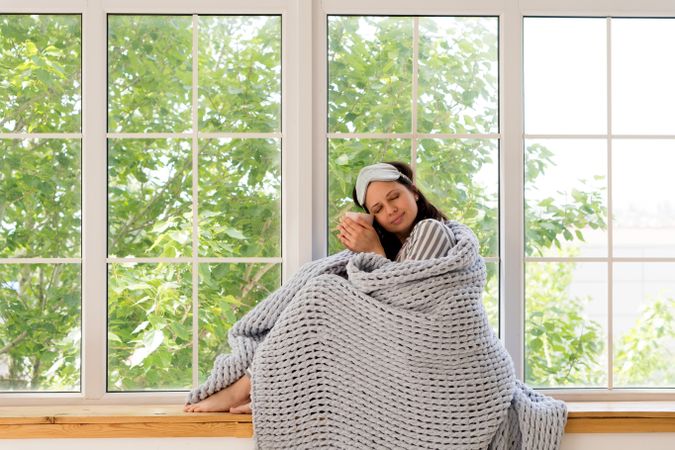 Woman enjoys tranquility, wrapped in gray blanket by window.