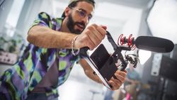 Young Male Filmmaker Operating A Modern Camera With Attached Microphone In A Studio, Capturing Content For A Creative Advertising Project, Showcasing Teamwork And Professional Equipment.