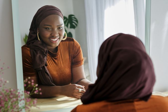 Happy Black female in hijab looking in mirror and smiling