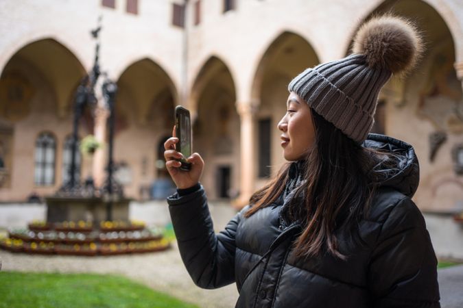 Content Asian female traveler in warm wear taking photo on smartphone while sightseeing at old city