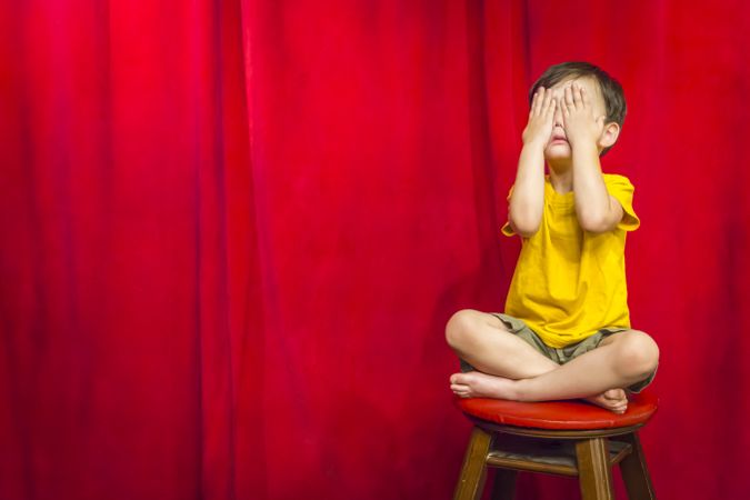 Boy Covering Eyes Sitting on Stool in Front of Curtain
