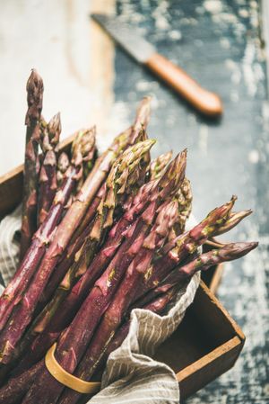 Close up of bunches of purple asparagus, knife on blue background, vertical composition