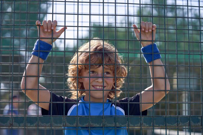 Cheerful boy hanging on metal fence on court on sunny day