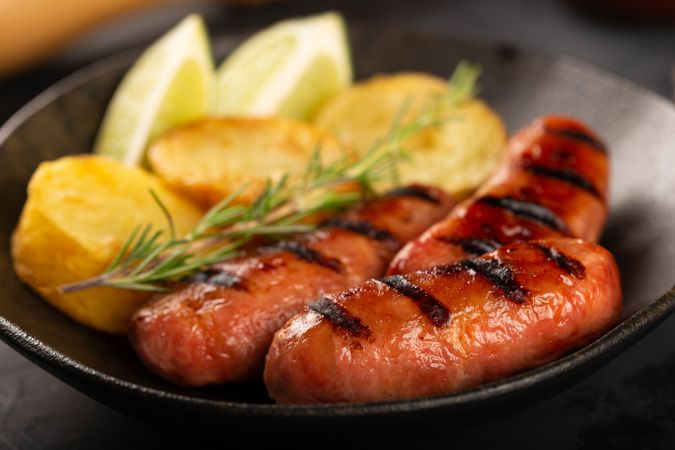 Close up of plate with grilled sausages, potatoes, lime slices and sprig of rosemary
