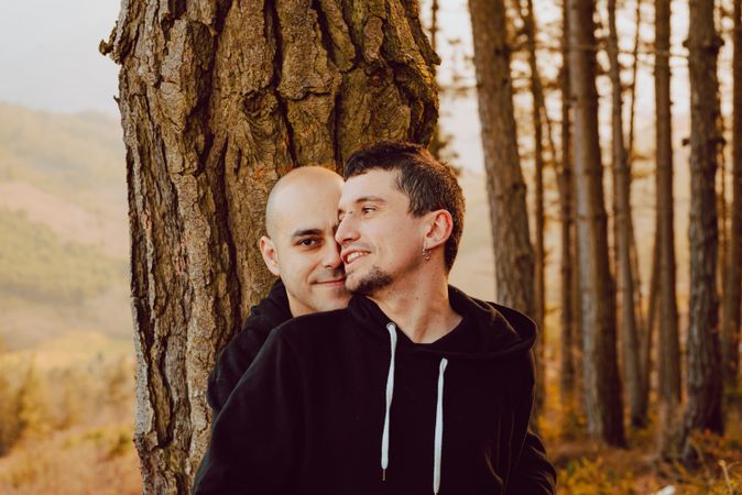 Smiling men hugging near tree in forest on hill