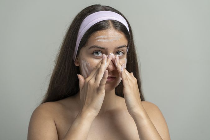 Beautiful woman looking at camera while washing her face with a cleansing scrub or mask
