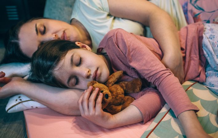 Portrait of serene daughter embracing a teddy bear lying next to mom over mats at night