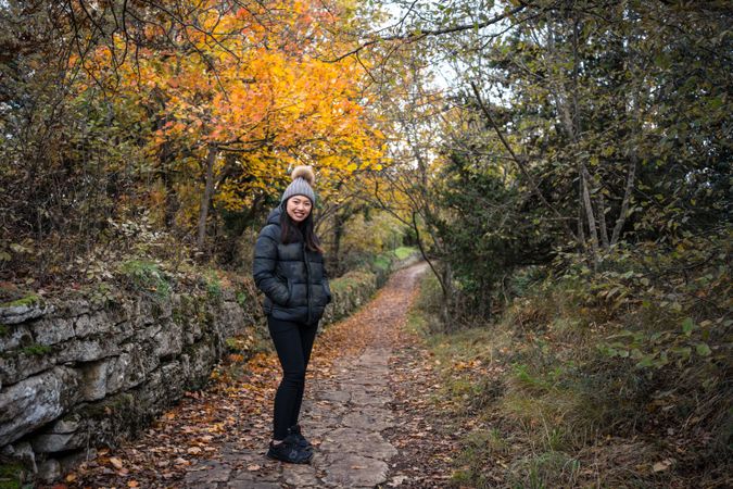 Smiling Asian woman in warm clothes standing in park with trees and stony fence