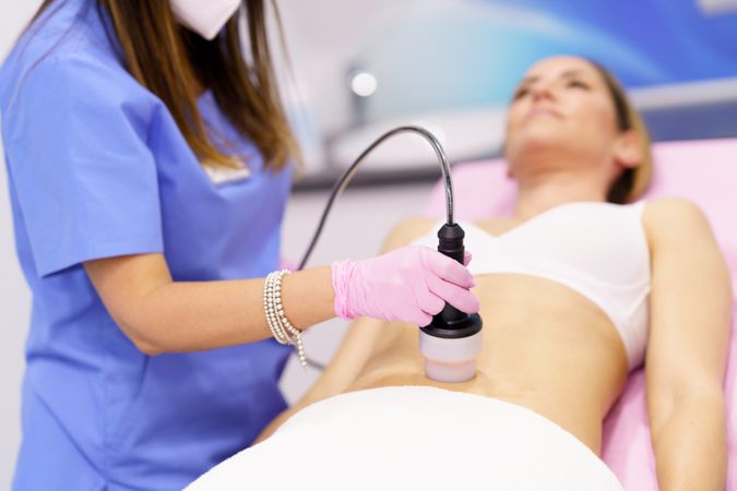 Professional in scrubs performing cosmetic procedure on woman’s stomach