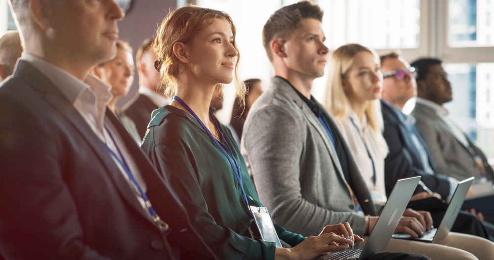 Young Woman Sitting in Crowded Audience at Business Conference. Female Delegate Smiling and Using Laptop Computer. Manager Watching Motivational Presentation About Investing In Innovative Startups.