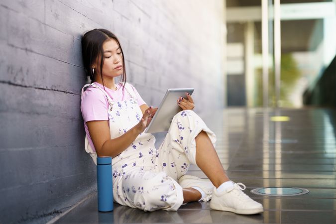 Woman sitting with digital tablet leaning against wall outside