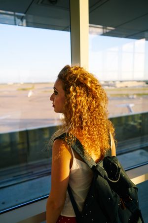 Young woman thoughtfully looking at airplanes through window in sunlight