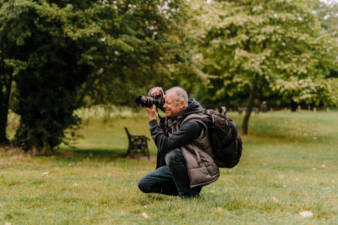 Older man crouching with large camera next to trees taking photos