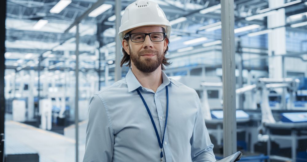 Young Electronics Manufacture Engineer Smiling, Looking at Camera. Portrait of a Man Wearing a Protective White Hard Hat. Specialist Monitoring Artificial Intelligence Systems at a Factory