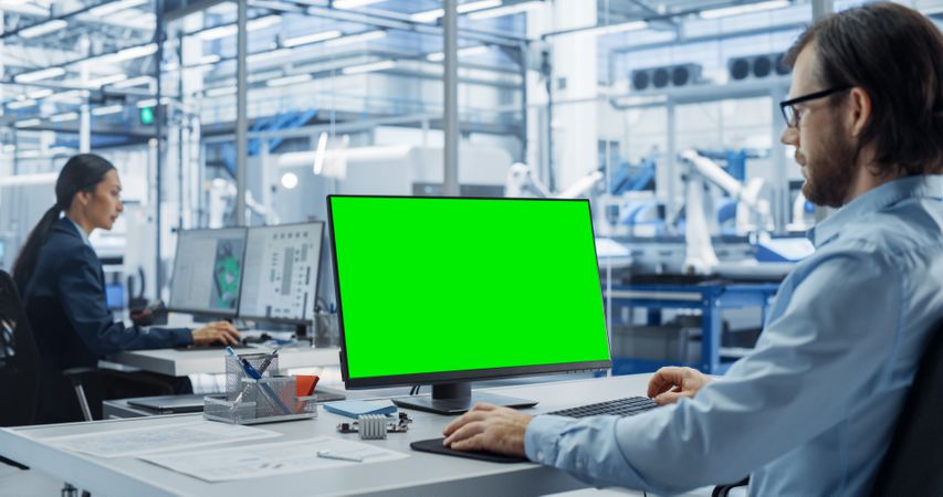Robotics Engineer Working on a Desktop Computer with Green Screen Chromakey Mock Up Display in a Factory Office. Technological Research and Development Facility with Autonomous Assembly Conveyor