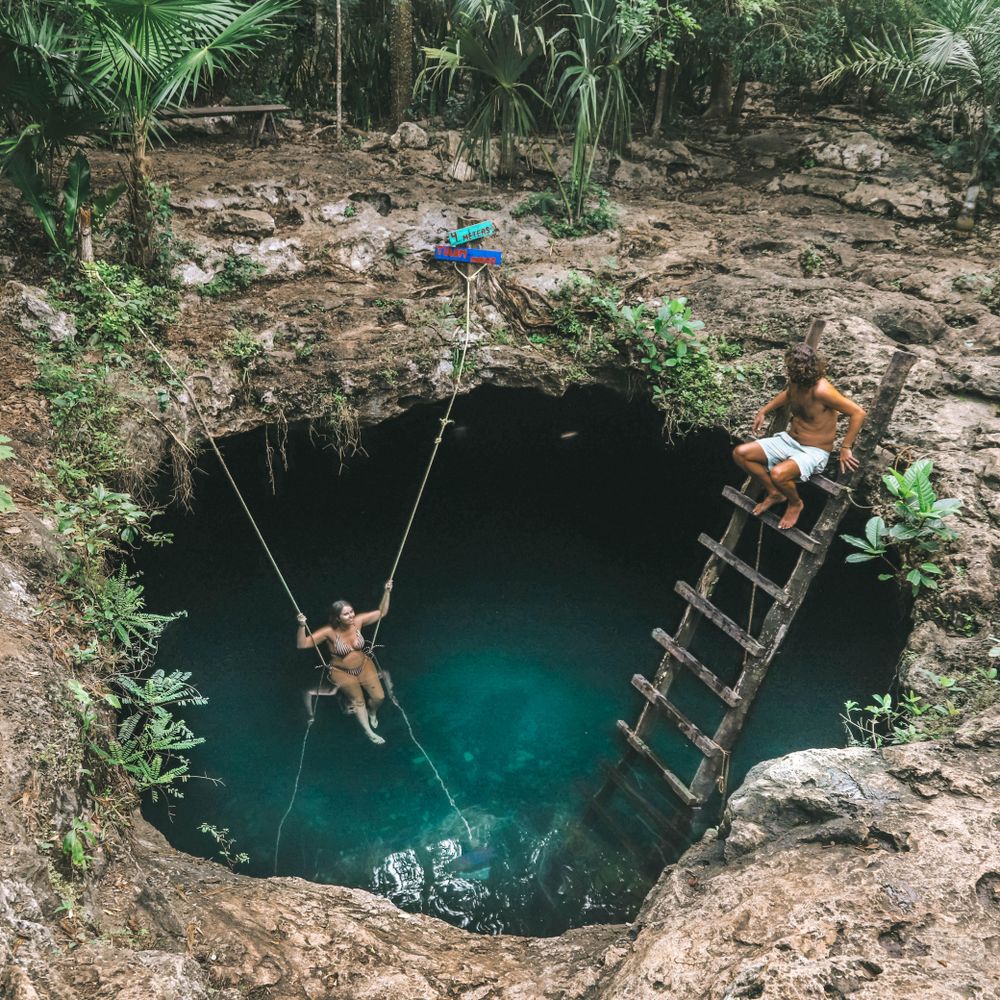 Woman on swing and man on wooden ladder above on an edge of a cave in forest