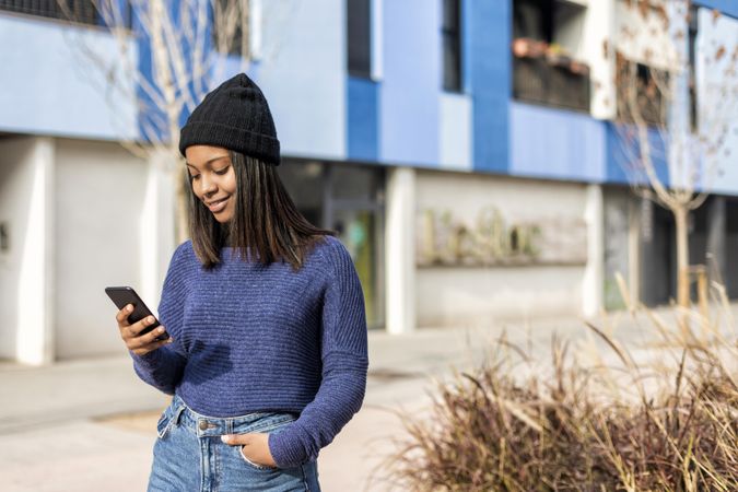 Female in wool hat and sweater checking phone outside blue building