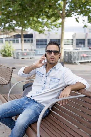 Male in denim shirt relaxing on city bench outside and speaking on phone