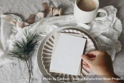 Hand placing blank card, invitation mockup on ceramic plate with pine branch on tablecloth 0LdEjV
